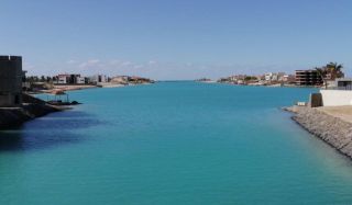 Fishing Yacht and Villa Rental on the Red Sea Package A serene turquoise waterway cuts through coastal buildings under a clear sky.