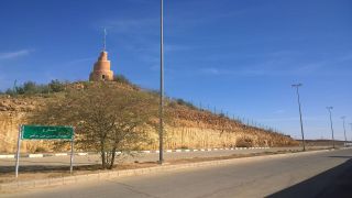 Al Majma’ah City Tour from Riyadh via SAR Train A desert road stretches alongside a rocky hill topped by a tower, under a clear blue sky. A green street sign stands beside the road.