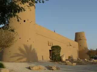 Central Riyadh Al Masmak Fort, Grand Mosque and Deira Souk Tour A tan-colored fortress with two prominent towers stands against a clear blue sky, with scattered rocks and greenery in the foreground.