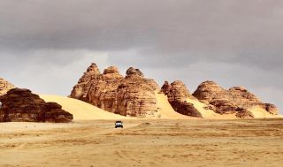 Saudi Arabia 13-Day Archaeological and Historical Tour A lone vehicle traverses a vast, sandy desert landscape dotted with unique rock formations under a cloudy sky.
