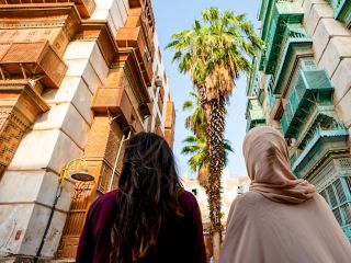 Saudi Arabia Museums, Roses and Souks: Jeddah and Taif 3-Day Tour Two women, one with dark hair and the other wearing a hijab, stand on a street lined with tall buildings, showcasing intricate woodwork and colorful balconies, under tall palm trees.