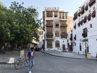 Jeddah Day Tour and Day Excursion from Jeddah – Old City and New City A man walks across a cobblestone street with his bicycle, surrounded by traditional buildings with wooden balconies and window coverings, with trees on the side.