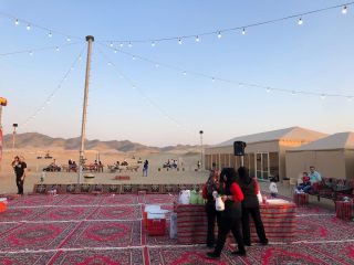 Group Day Desert Luxury Camp Excursion and Off-Road Driving from Jeddah An outdoor desert gathering features tents, string lights, and people socializing, with some seated and others being served by women in black and red attire.