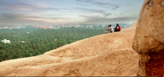Al Ahsa Mountains and Green Desert Oasis Three people sit atop a rocky outcrop, overlooking a dense forest and a distant cityscape under a cloudy sky.