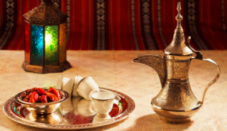 Jeddah Food, Coffee, Tea, and Snacks Tour A table is set with dates, small cups, and a traditional Arabic coffee pot, illuminated by a colorful lantern, against a red patterned backdrop, suggesting a cultural or festive occasion.