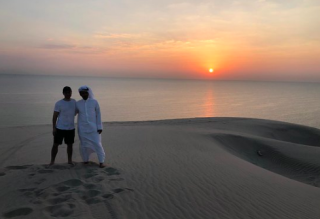 Saudi Seaside & Shoreside Tour: Persian Gulf Explorer (From Dammam/Al Khobar) Two men, one in western attire and the other in traditional Arab clothing, stand on a sand dune near the sea during a vibrant sunset.
