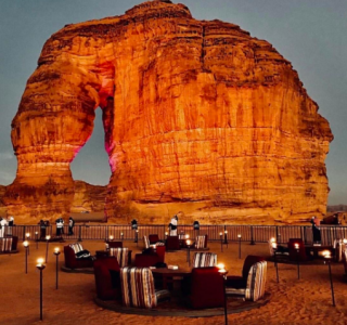 Saudi-Cross Tour: From the Persian Gulf to the Red Sea Total Tour A giant sandstone rock formation with an arch stands majestically behind an outdoor seating area with chairs, tables, and small lights.