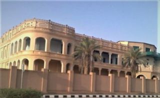 Al Kharj Museum, Palace and Tomb Day Tour from Riyadh A beige building with arched openings and a surrounding wall sits under a clear blue sky, with palm trees in front of it.