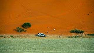 Al Kharrarah National Park Desert Adventure from Riyadh An aerial view of an SUV parked on a sandy area between a green field and an orange sand dune with people walking around.