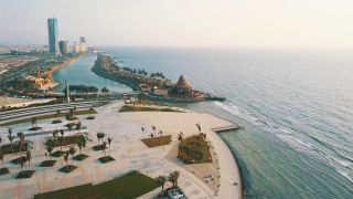Saudi Arabia Ethnic Trip via Jeddah, Medina & AlUla Aerial shot of a coastal cityscape featuring modern buildings, a mosque, a pier with a unique conical-roofed structure, and a park with palm trees, along a curved shoreline.