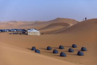 5-Day Hijrah Route Tour A desert landscape features a campsite with tents, vehicles, and people, set against a backdrop of rolling sand dunes and a clear sky.
