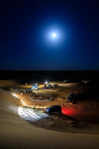 Makkah to Madinah Trekking Tour (3 weeks) A group of people gathers in the desert at night, illuminated by vehicle headlights and a bright moon.